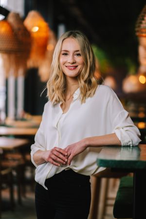 Eine lächelnde Frau mit blondem Haar, die eine weiße Bluse und eine schwarze Hose trägt, steht in einem stilvollen, warm beleuchteten Restaurant mit Holztischen und Hängelampen im Hintergrund.