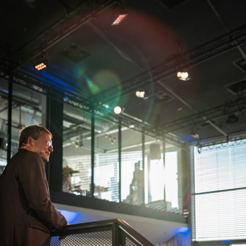 Ein Mann im Anzug steht auf einer Treppe in einem modernen Gebäude mit großen Fenstern und hellem Licht, das hereinströmt, und schaut nachdenklich hinaus.