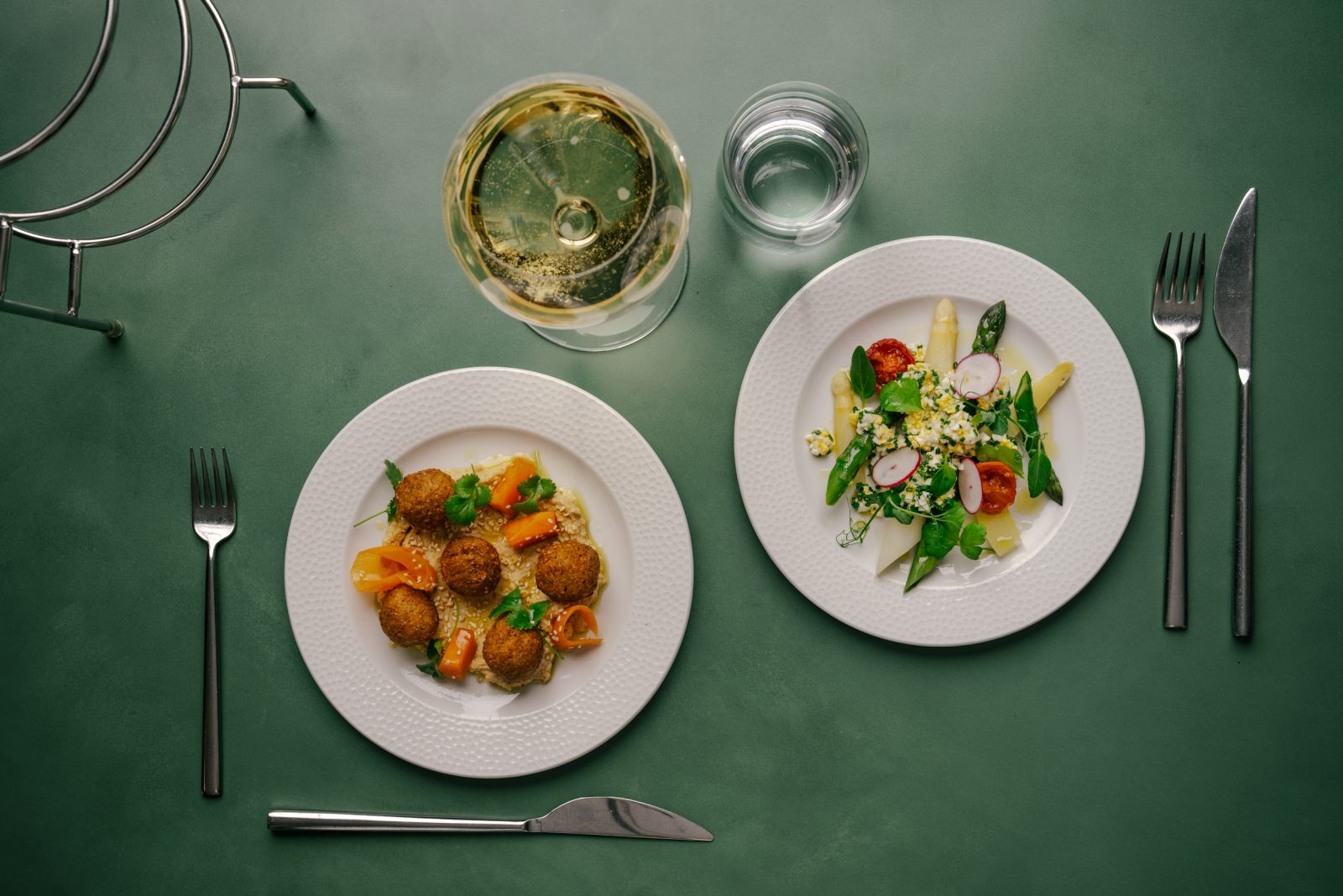 Zwei weiße Teller auf einem grünen Tisch: auf dem einen liegen Falafel und Orangenscheiben, auf dem anderen ein frischer Salat, daneben ein Glas Weißwein, ein Glas Wasser und Besteck, das auf beiden Seiten fein säuberlich angeordnet ist.