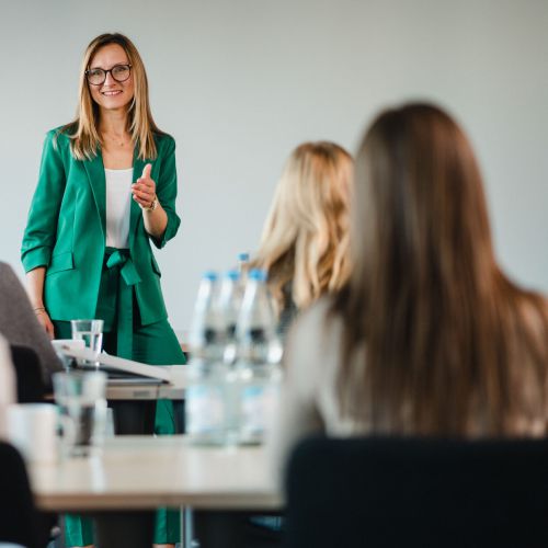 Eine Frau in einem grünen Anzug steht auf und spricht zu einer Gruppe von Menschen, die an einem Tisch sitzen und eine Präsentation in einem hellen, modernen Konferenzraum halten.