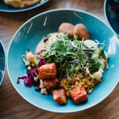 Eine blaue Schale, gefüllt mit farbenfrohem geröstetem Gemüse, Körnern, Mikrogrün, Falafelbällchen und Tofu- oder Paneerwürfeln, angeordnet auf einem Holztisch mit anderen ähnlichen Schalen im Hintergrund.