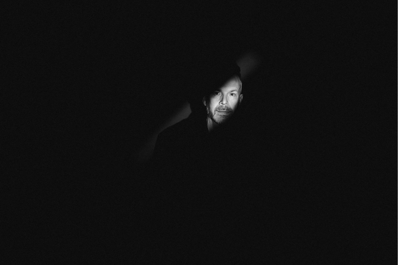 Das Gesicht eines Mannes wird in einer dunklen, schattigen Umgebung teilweise beleuchtet, wobei der größte Teil des Bildes in tiefem Schwarz gehalten ist und nur ein kleiner Teil des Gesichts in einem Lichtstrahl sichtbar ist.