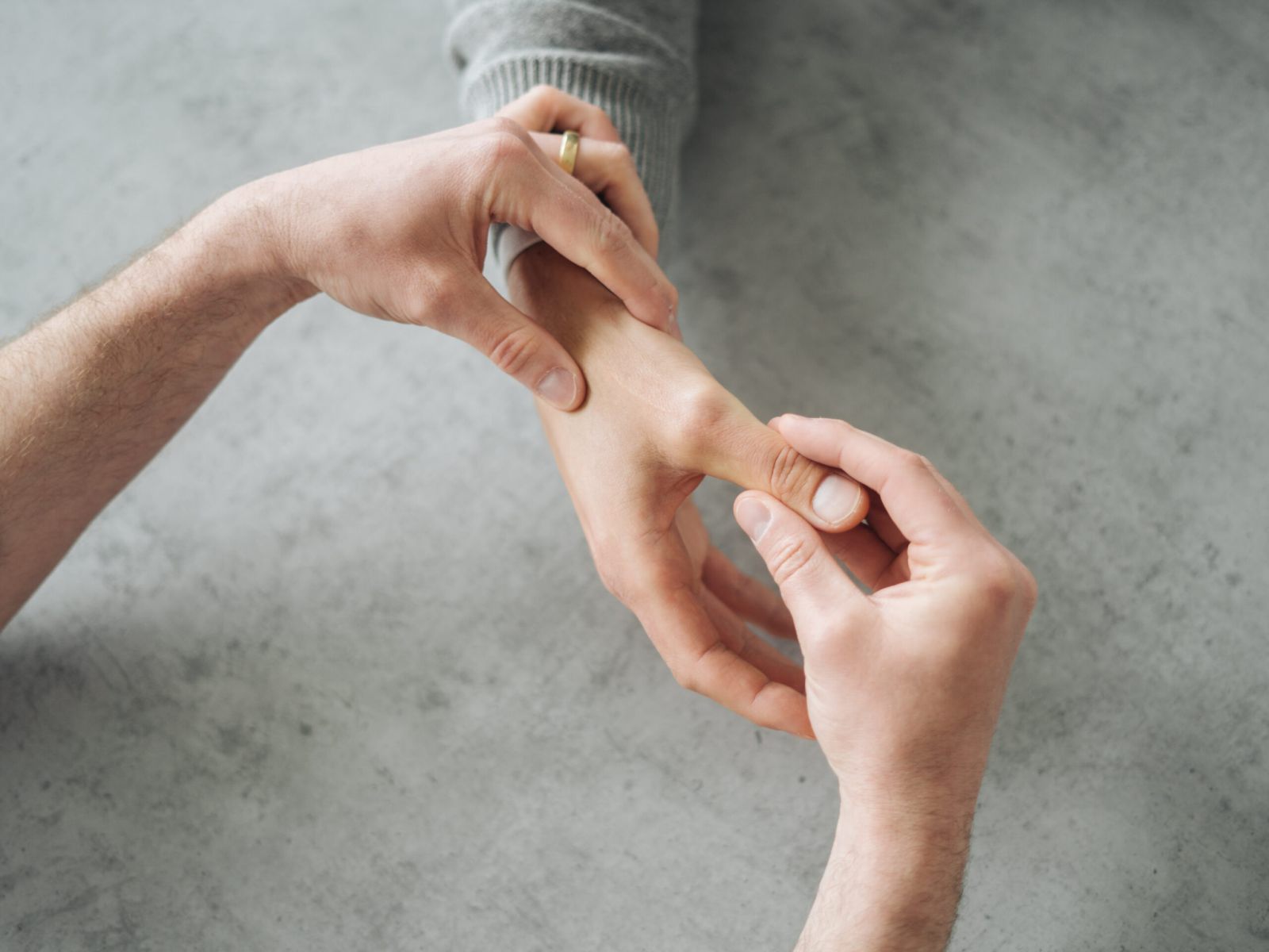Eine Nahaufnahme einer Person, die mit beiden Händen die Daumenwurzel einer anderen Person massiert, möglicherweise zur Schmerzlinderung oder zur physikalischen Therapie, auf einer hellgrauen Fläche.