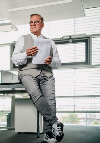 Ein Mann mit Brille, weißem Hemd und grauer Weste sitzt auf der Kante eines Schreibtisches in einem modernen Büro, hält Papiere in der Hand und schaut nachdenklich zur Seite. Im Hintergrund stehen ein leerer Stuhl und ein Computer.
