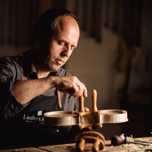 Ein Mann in einer Werkstatt, der sorgfältig ein hölzernes Streichinstrument, möglicherweise eine Geige, mit Handwerkzeugen schnitzt oder zusammenbaut. Die Szene ist warm beleuchtet und konzentriert sich auf die Handwerkskunst und die Konzentration.