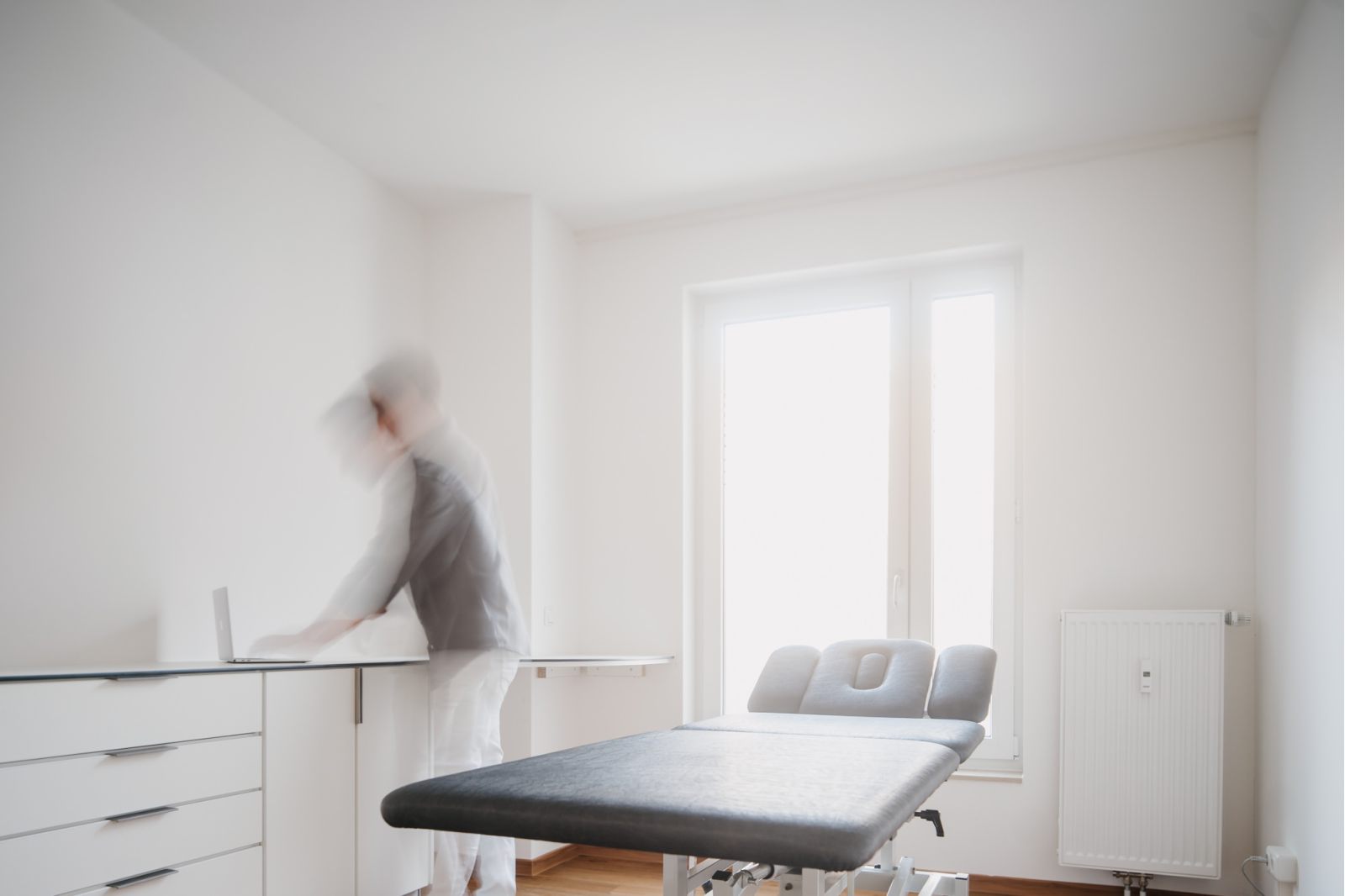 Ein heller, minimalistischer medizinischer Untersuchungsraum mit einem Behandlungstisch, weißen Wänden und einem Fenster. Eine verschwommene Person steht in der Nähe eines Tresens und suggeriert Bewegung. Der Raum wirkt sauber und modern.