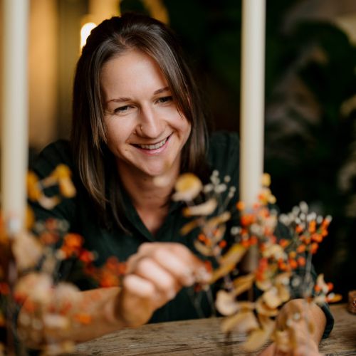 Eine lächelnde Frau arrangiert herbstliche Zweige mit orangefarbenen Beeren und getrockneten Blättern auf einem Holztisch, daneben stehen zwei hohe weiße Kerzen in Haltern. Der Hintergrund ist sanft beleuchtet und unscharf.
