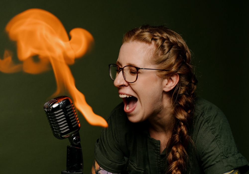 Eine Frau mit Brille und geflochtenem Haar, die bunte Ärmeltätowierungen zeigt, lehnt sich an ein altes Mikrofon und scheint zu schreien, während eine orangefarbene Flamme aus dem Mikrofon vor einem grünen Hintergrund aufsteigt.
