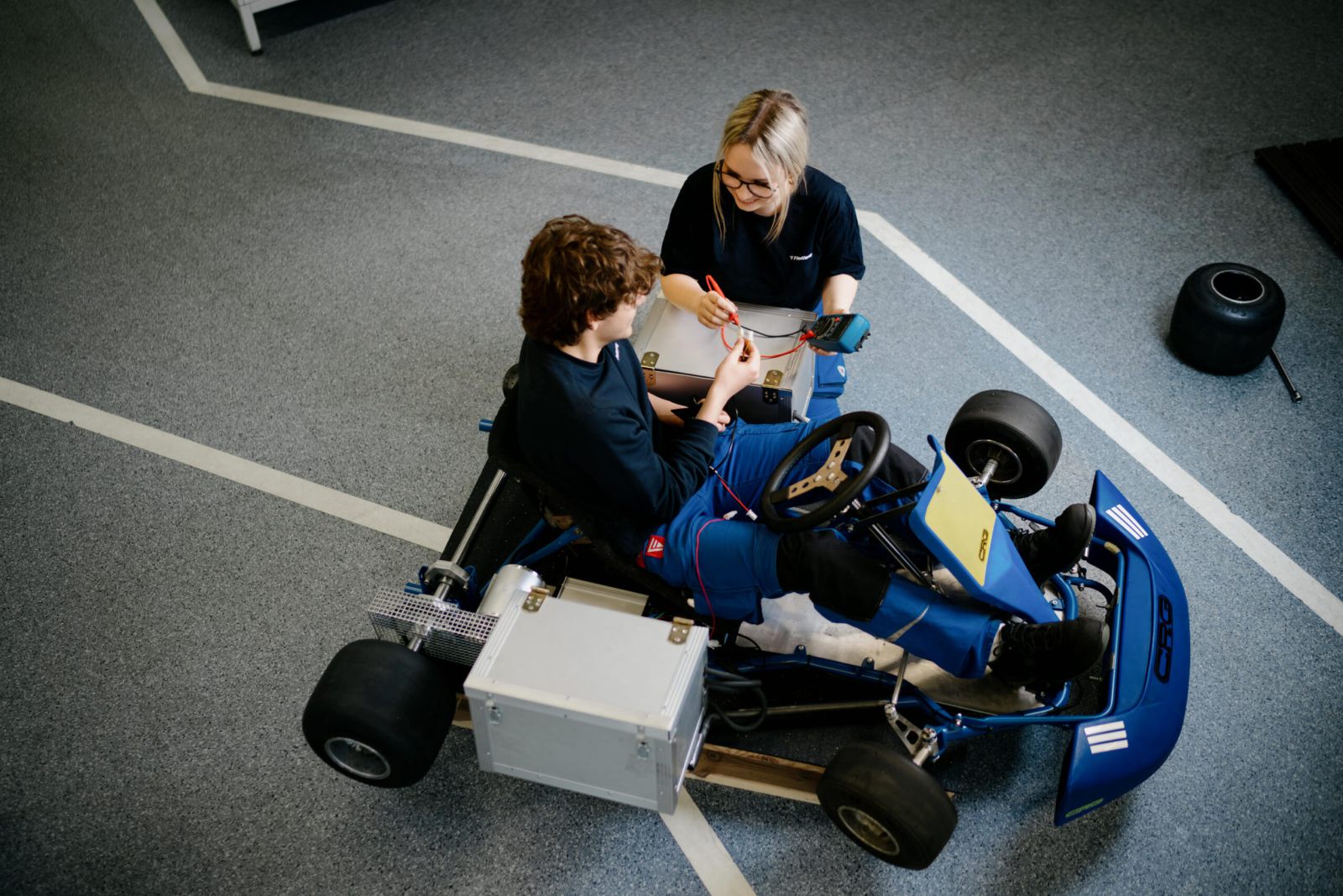 Eine Person, die in einem Go-Kart sitzt, wird von einer anderen Person unterstützt, die Kabel und Werkzeuge in der Hand hält und möglicherweise Wartungs- oder Reparaturarbeiten durchführt. Die Szene spielt in einer Halle auf einer markierten Strecke.