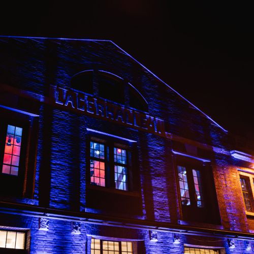Ein Backsteingebäude, das nachts mit blauen und orangefarbenen Lichtern beleuchtet wird. Der Schriftzug "LAGERHALLE" ist über den Fenstern zu sehen, die von innen in rotem und blauem Licht leuchten.