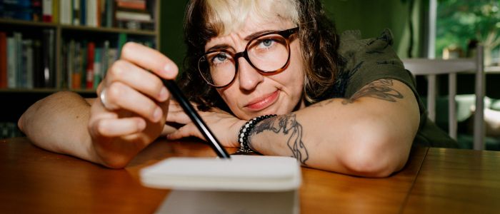 Eine Person mit Brille und gewelltem, teilweise blondem Haar stützt ihren Kopf auf einen Holztisch, hält einen Stift über ein aufgeschlagenes Notizbuch und schaut nachdenklich. Im Hintergrund sind Bücherregale und Pflanzen zu sehen.