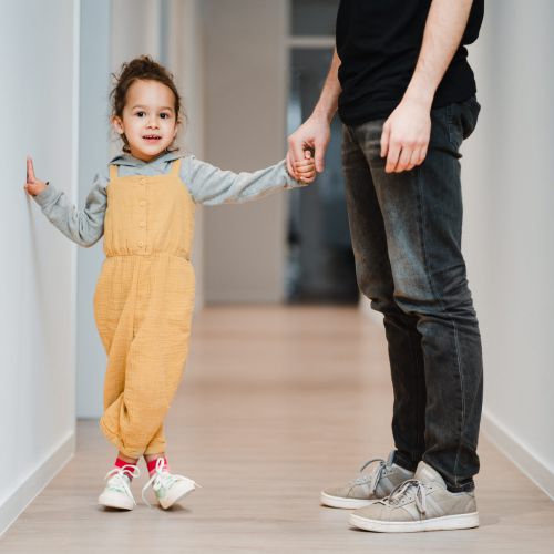 Ein kleines Kind in gelber Latzhose hält sich an der Hand eines Erwachsenen in Jeans, der einen hellen Flur entlanggeht. Das Kind lächelt und berührt mit einer Hand die Wand des Flurs.