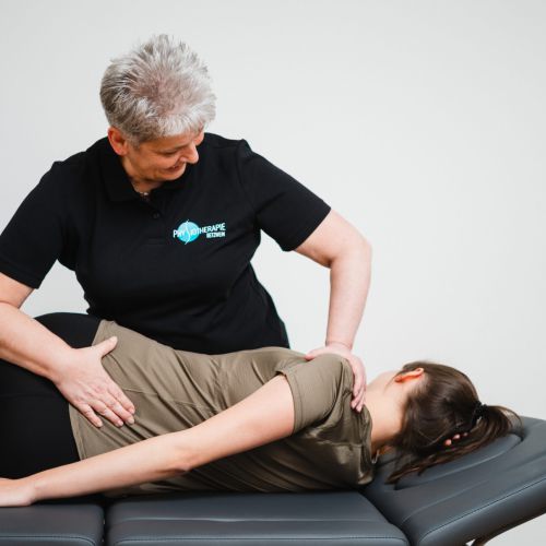Ein Physiotherapeut in einem schwarzen Polohemd hilft einer Frau, die auf der Seite auf einer Behandlungsliege liegt, und dehnt sanft ihren Rücken und ihre Hüfte in einer klinischen Umgebung.