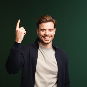 Ein lächelnder Mann mit braunem Haar, hellem Hemd und dunkler Jacke steht vor einem grünen Hintergrund und hebt den rechten Zeigefinger, als ob er etwas sagen oder eine Idee haben würde.