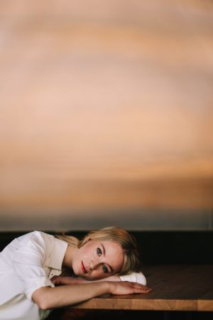 Eine Frau mit blondem Haar, die ein weißes Hemd trägt, stützt ihren Kopf und ihre Arme auf einen Holztisch und blickt nachdenklich nach vorn. Der Hintergrund ist in warmen, erdigen Tönen sanft verschwommen.