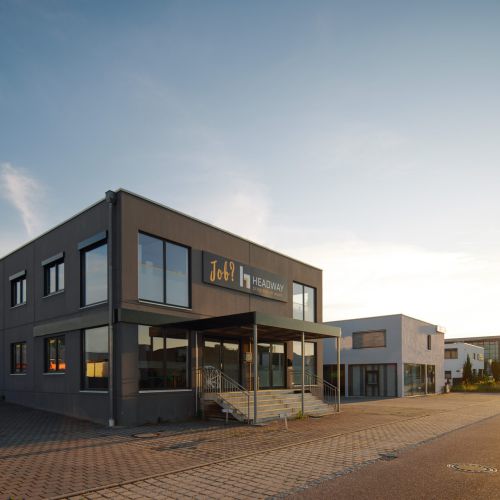 Modernes graues Industriegebäude mit großen Fenstern und einem Schild mit der Aufschrift "Job7 HEADWAY" über dem Eingang, gelegen an einer gepflasterten Straße unter einem klaren Himmel bei Sonnenuntergang.