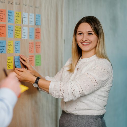 Eine Frau lächelt und klebt eine Haftnotiz auf eine mit bunten Zetteln bedeckte Tafel, während ein Mann mit einem Notizblock in der Hand daneben steht, beide sind in eine gemeinsame Planungssitzung vertieft.