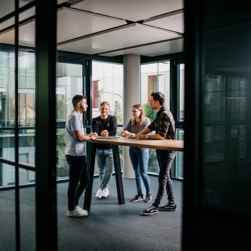 Vier junge Erwachsene stehen um einen hohen Tisch in einem modernen Büro mit Glaswänden, unterhalten sich und lächeln. Der Raum ist von natürlichem Licht durchflutet, und vor den Fenstern sind die Gebäude der Stadt zu sehen.