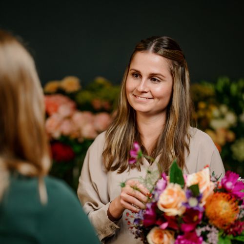 Eine Frau, die einen bunten Blumenstrauß in der Hand hält, lächelt und unterhält sich mit einer anderen Person in einem Blumenladen, wobei verschiedene Blumen im Hintergrund verschwimmen.