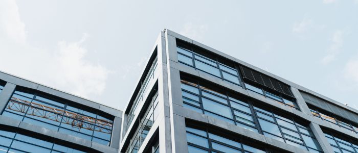 Modernes Bürogebäude mit großen Glasfenstern und einem Betonrahmen, aus einem niedrigen Winkel gegen einen teilweise bewölkten Himmel gesehen. Ein Baum und ein Baukran sind auf der linken Seite teilweise sichtbar.