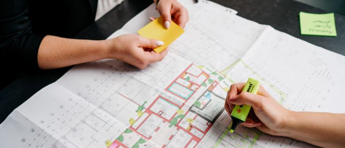 Zwei Personen betrachten einen bunten Gebäudegrundriss, der auf einem Tisch ausgebreitet ist. Eine Person hält gelbe Haftnotizen in der Hand, die andere hebt Abschnitte des Plans mit einem grünen Textmarker hervor.