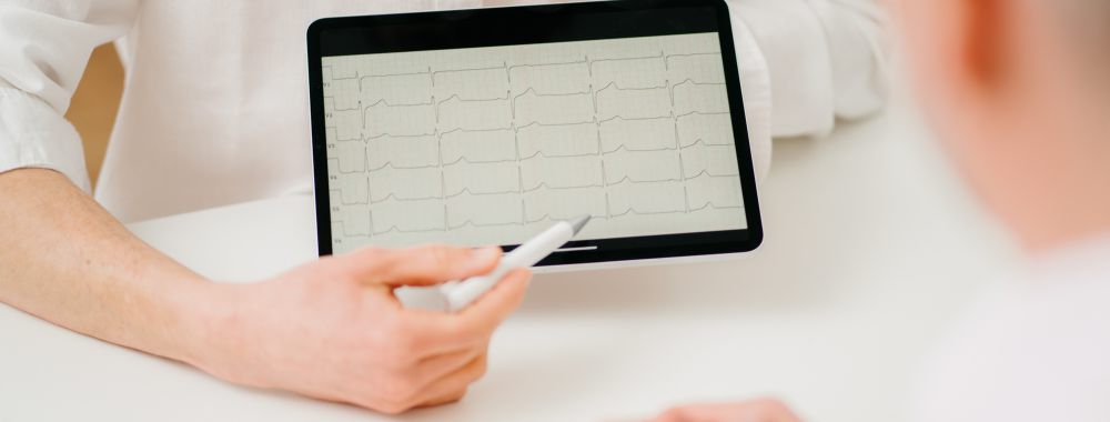 Eine Person in einem weißen Hemd zeigt mit einem Stift auf ein Elektrokardiogramm (EKG), das auf einem Tablet angezeigt wird, während eine andere Person am anderen Ende des Tisches aufmerksam zuhört.