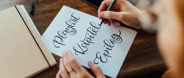 Eine Person übt sich in moderner Kalligrafie und schreibt die Worte Playlist, Kapiteln und Epilog kursiv auf weißem Papier an einem hölzernen Schreibtisch mit einem Notizbuch, einem Stift und einem Tablet in der Nähe.