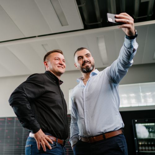 Zwei Männer stehen Seite an Seite, lächeln und machen ein Selfie in einem modernen Büro. Im Hintergrund unterhalten sich ein Mann und eine Frau in der Nähe der Fenster. Die Umgebung ist hell und professionell.