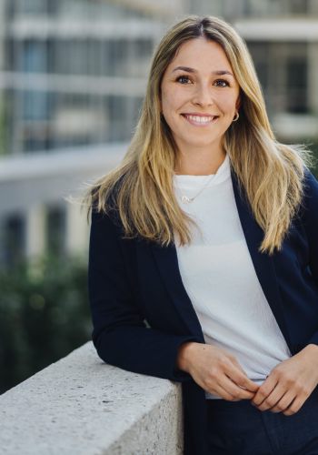 Eine Frau mit langen blonden Haaren, die einen marineblauen Blazer und ein weißes Hemd trägt, lächelt, während sie sich auf einen Betonsims im Freien lehnt, mit modernen Gebäuden und Grünflächen im Hintergrund.