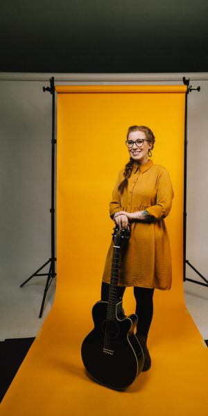 Eine Frau mit Brille und rotem Haar steht lächelnd vor einem gelben Hintergrund, trägt ein gelbes Kleid und braune Stiefel und stützt ihre Hände auf den Hals einer schwarzen Akustikgitarre.