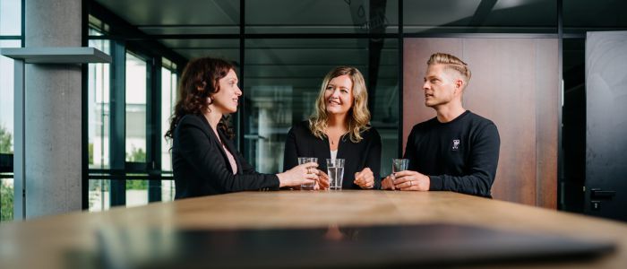 Drei Personen stehen in einem modernen Büro um einen Tisch herum, halten Gläser mit Wasser in der Hand und lächeln, während sie ein Gespräch führen. Im Hintergrund sind große Fenster und Glaswände zu sehen.