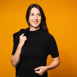 Eine Frau mit langen braunen Haaren steht vor einem orangefarbenen Hintergrund, lächelt und blickt zur Seite. Sie trägt ein schwarzes Hemd und hat eine Jacke über die Schulter gehängt.