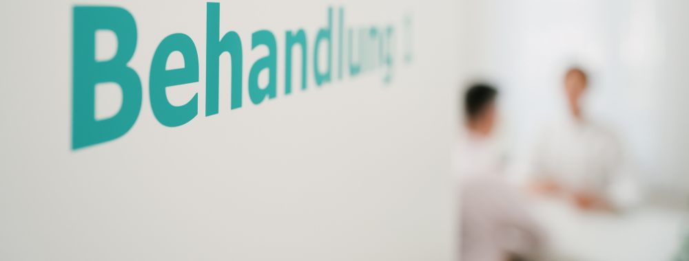 Eine weiße Wand mit dem Wort "Behandlung" in blaugrüner Schrift; im Hintergrund sind zwei Personen in weißer Kleidung zu sehen, die in einer hellen, klinischen Umgebung sitzen und sprechen.