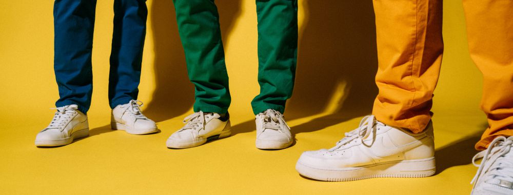 Drei Personen stehen vor einem gelben Hintergrund und tragen bunte Hosen - blau, grün und gelb - sowie weiße Turnschuhe. Nur ihre Beine und Füße sind sichtbar.