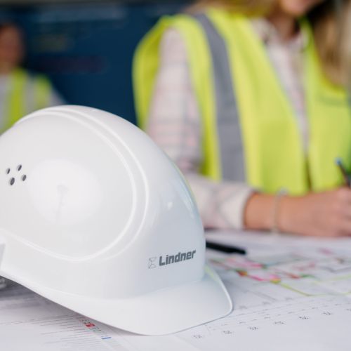 Ein weißer Lindner-Schutzhelm ruht auf Bauunterlagen, während eine Person in einer gelben Sicherheitsweste im Hintergrund auf Papieren schreibt. Die Szenerie lässt auf eine Bau- oder Projektplanungsumgebung schließen.