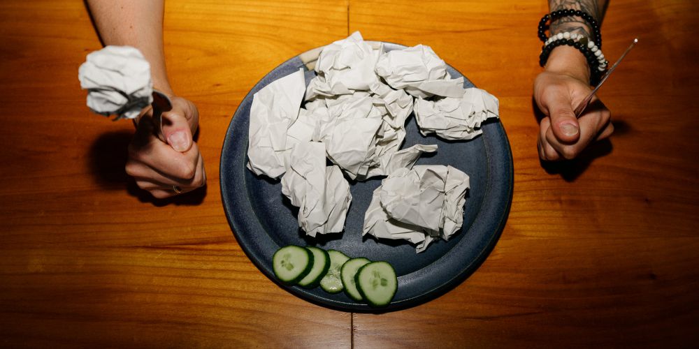 Eine Person sitzt an einem Holztisch und hält Gabel und Messer über einen Teller mit zerknülltem Papier und ein paar Gurkenscheiben. Nur ihr Oberkörper und ihre Arme sind sichtbar.