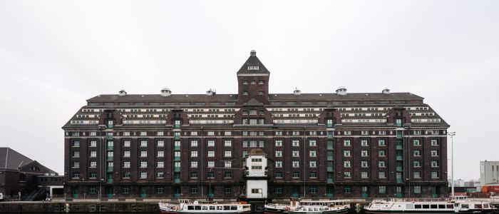 Ein großes, dunkles Backsteingebäude mit einem zentralen Turm steht am Wasser, mit mehreren weißen Booten am Rand und einem Kopfsteinpflasterweg im Vordergrund unter einem bedeckten Himmel.