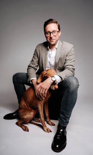 Ein Mann mit Brille, grauem Blazer, weißem Hemd und Jeans sitzt lächelnd auf einem Hocker, eine Hand stützt sich auf einen braunen Hund, der dicht neben seinen Beinen sitzt. Der Hintergrund ist schlicht und in hellen Farben gehalten.