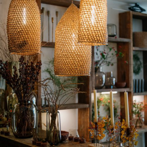 Drei geflochtene Pendelleuchten hängen über einem rustikalen Holztisch, der mit Glasvasen mit getrockneten Blumen, Kerzen und kleinen Dekorationsgegenständen dekoriert ist, in einem gemütlichen, warm beleuchteten Raum mit Holzregalen und Pflanzen im Hintergrund.