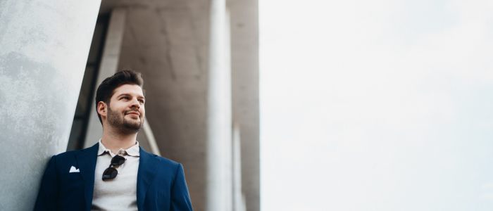 Ein Mann in blauem Anzug und weißem Hemd steht im Freien, lehnt an einem Betonpfeiler eines modernen Gebäudes und blickt mit einem leichten Lächeln zur Seite. An seinem Hemdkragen hängt eine Sonnenbrille. Der Himmel ist klar.