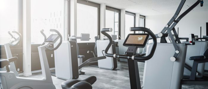 Ein modernes Fitnessstudio mit großen Fenstern und natürlichem Licht, das mit verschiedenen Fitnessgeräten, darunter stationäre Fahrräder und Krafttrainingsgeräte, auf einem sauberen, grauen Boden ausgestattet ist.