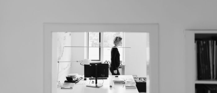 Ein Schwarz-Weiß-Foto zeigt eine Frau, die in einem modernen Büro in der Nähe eines Schreibtisches mit einem Computer und Büchern steht, gesehen durch eine Tür. Die Szene ist minimalistisch und durch natürliches Licht gut beleuchtet.