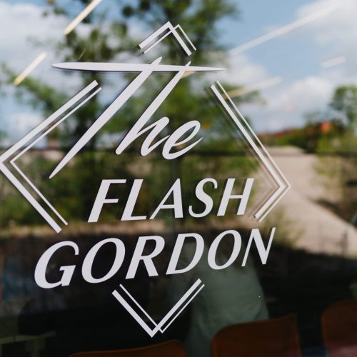 Der weiße Text auf einem Glasfenster lautet "The Flash Gordon" in fetten, stilisierten Buchstaben mit dekorativen Rautenformen um die Worte herum; in der Reflexion sind Bäume und der Himmel zu sehen.