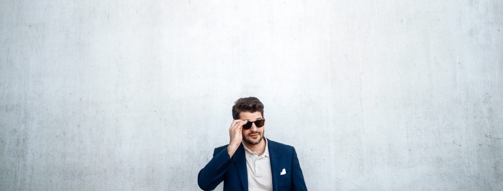 Ein Mann im blauen Anzug und mit Sonnenbrille steht vor einer großen, schlichten hellgrauen Betonwand und rückt mit der Hand seine Sonnenbrille zurecht. Der Mann wirkt klein im Vergleich zu dem riesigen leeren Raum über ihm.