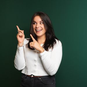 Eine Frau mit langen dunklen Haaren, die ein weißes Hemd und eine schwarze Hose trägt, lächelt und zeigt mit beiden Händen nach oben vor einem schlichten dunkelgrünen Hintergrund.