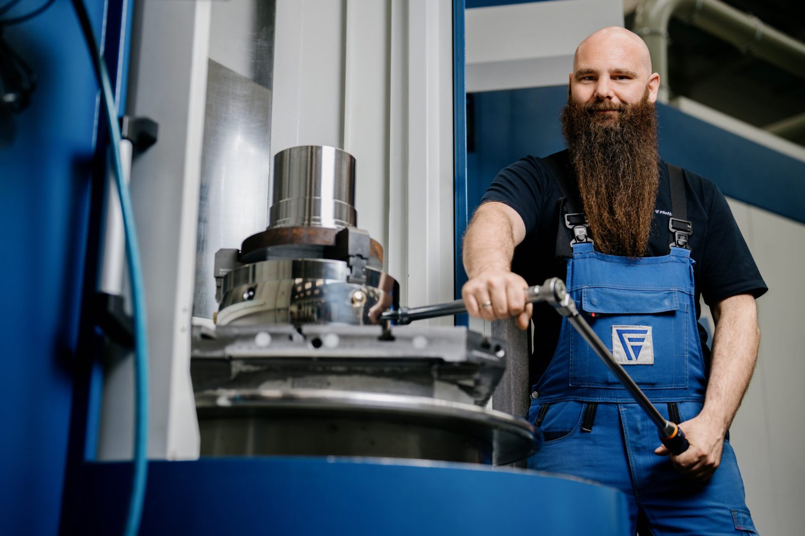Ein Mann mit langem Bart, der einen blauen Overall trägt, bedient in einer Fabrikhalle Industriemaschinen und lächelt in die Kamera, während er ein Werkzeug hält.