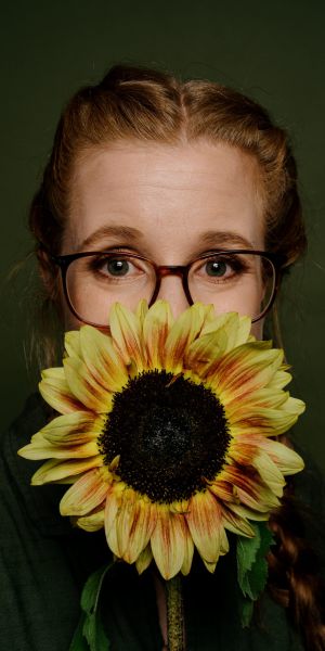 Eine Frau mit Brille und geflochtenem Haar hält sich eine große Sonnenblume vor Mund und Nase, so dass nur ihre Augen und ihre Stirn zu sehen sind. Der Hintergrund ist einfarbig dunkelgrün.
