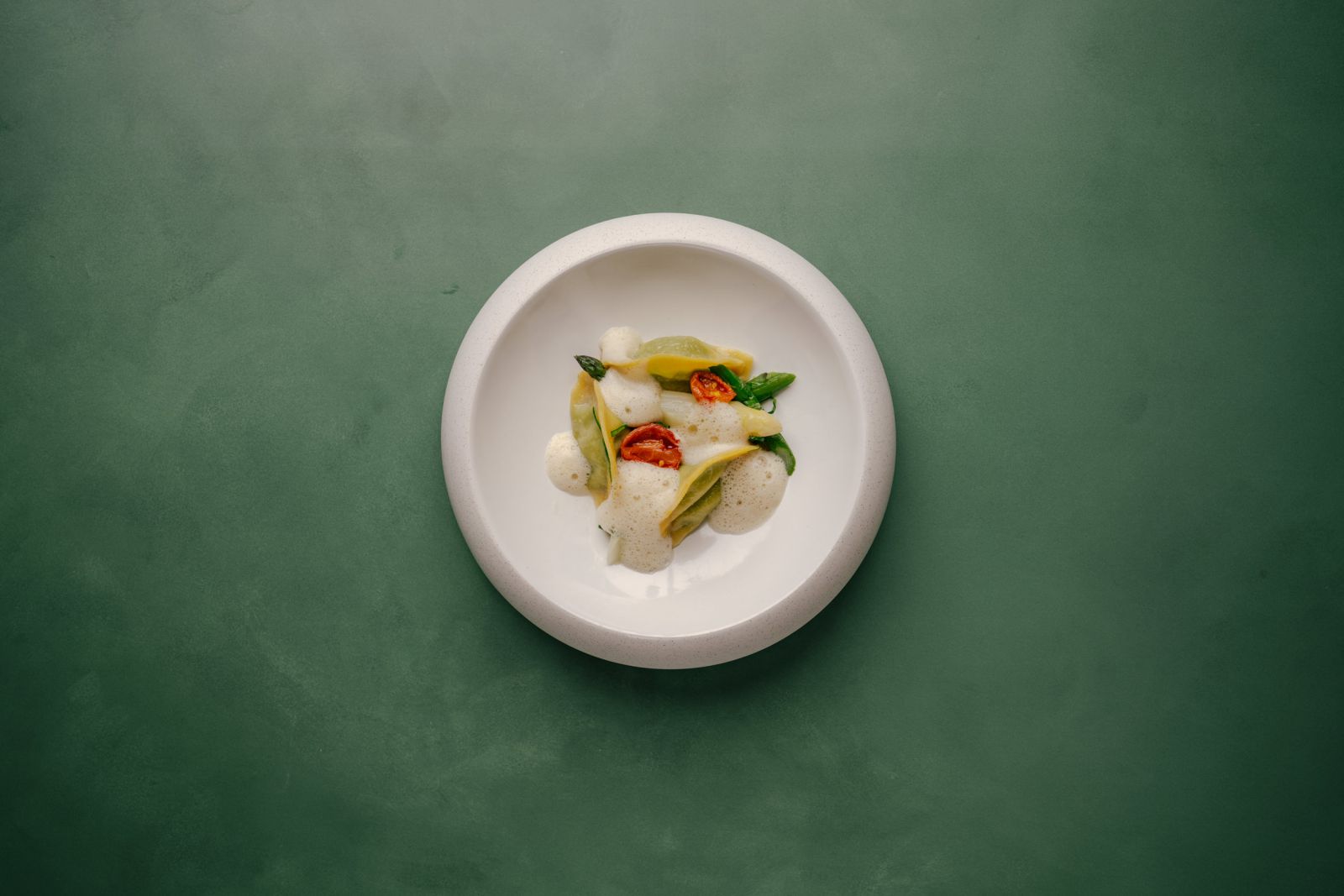 Ein weißer Teller mit einer kleinen Portion Gourmet-Pasta, garniert mit Schaum, Spargel und Kirschtomatenhälften, auf einer grünen Fläche.