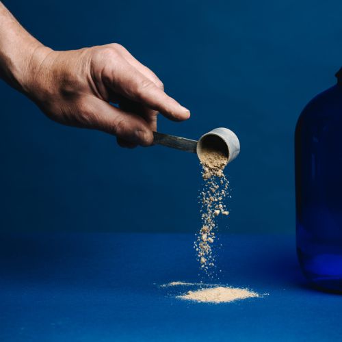 Eine Hand hält einen Messlöffel aus Metall und schüttet eine pulverförmige Substanz auf eine blaue Fläche, wobei rechts ein großer blauer Behälter vor blauem Hintergrund teilweise sichtbar ist.