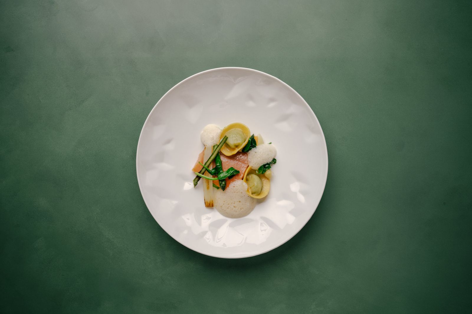 Ein Gourmet-Gericht mit kunstvoll arrangiertem Gemüse, Grünzeug und leichter Soße wird auf einem weißen, strukturierten Teller auf grünem Hintergrund serviert. Die Präsentation ist minimalistisch und elegant.